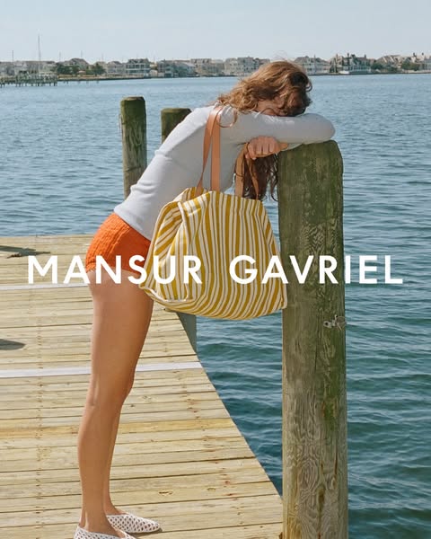 This Mansur Gavriel ad shows a model leaning on a wooden pier with the brand's tote bag hanging from her shoulder. T