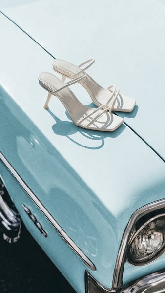 A pair of white high-heeled sandals are displayed on the hood of a baby blue car.