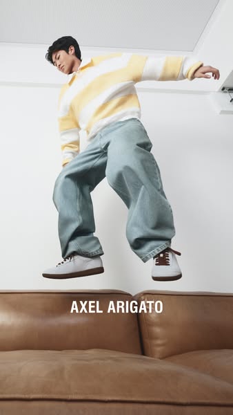 An image ad featuring a model in Axel Arigato sneakers, jeans, and a striped rugby shirt jumping over a brown leather co