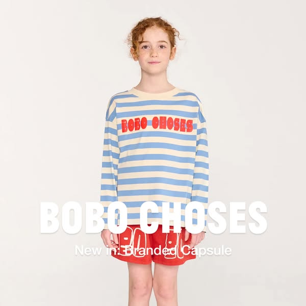 A girl is wearing a long sleeve shirt with blue and white stripes and red shorts. The clothing is from Bobo Choses.