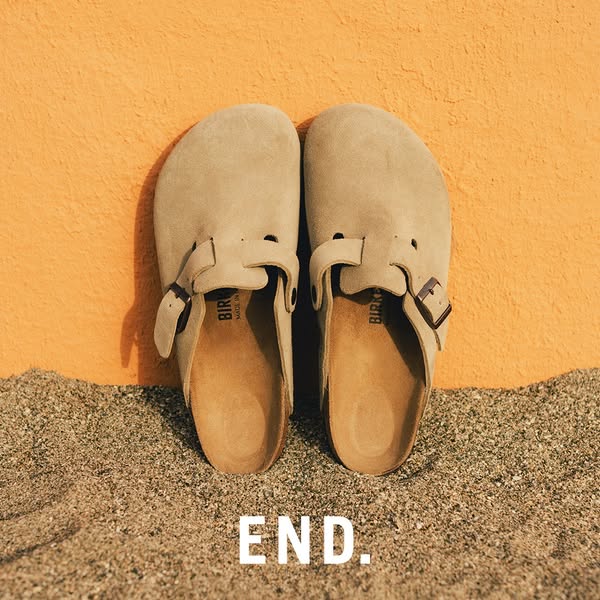 This image ad features two suede Birkenstock clogs resting on sand in front of an orange wall. The brand name is printed