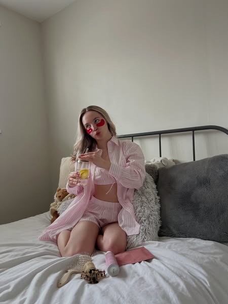 An image ad featuring a woman in a pink outfit sitting on a bed with products around her. She is holding a drink and wea