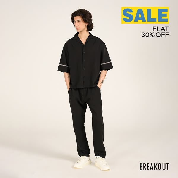An ad for Breakout clothing. It features a male model wearing a black two piece set and white sneakers against a light b