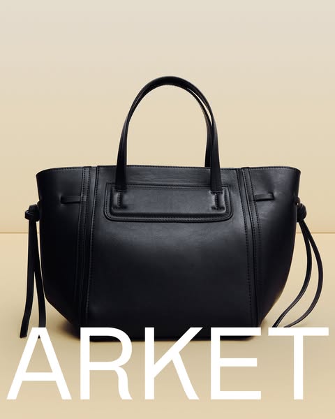 This is a minimalist product shot showcasing a black leather tote bag against a light beige background. The bag is the c