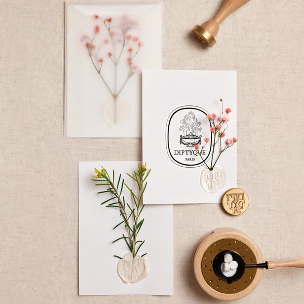 This image ad features a flatlay of Diptyque product packaging with wax seal stamps and flowers, showcasing the brand’s