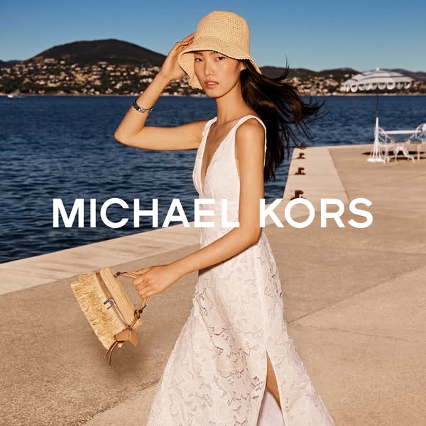 This ad showcases a woman walking along a seaside boardwalk wearing a dress, hat, and carrying a purse from Michael Kors