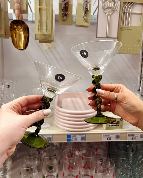 A pair of hands holds two martini glasses with dark green stems. The glasses are set in front of a shelf in a store with