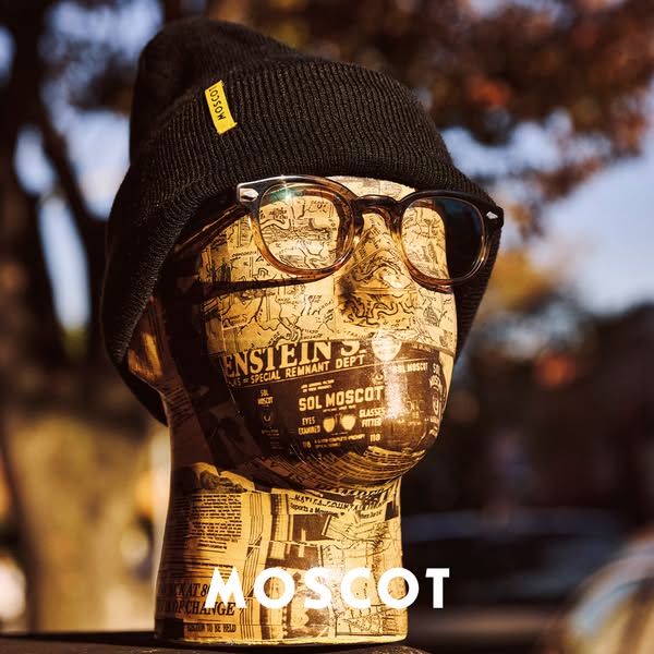 An ad featuring Moscot glasses on a head model with newspaper print paired with a black beanie. The Moscot brand is disp