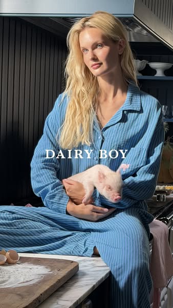 This image features a blonde woman in blue pajamas, sitting on a kitchen counter, and holding a small piglet.