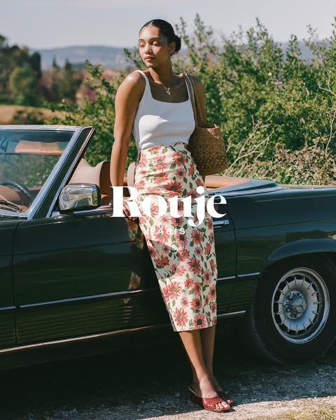 An ad showing a woman leaning against a car while wearing a skirt and top from the brand Rouje.