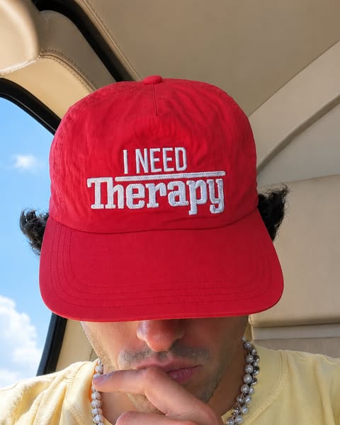 A close up image of a person wearing a red baseball cap with the phrase "I NEED Therapy" in white font. This a