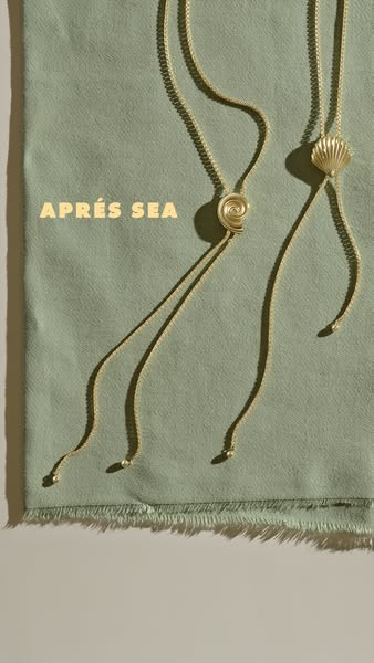 An ad for APrés Sea jewelry, featuring a still-life image of two gold necklaces against a green textile backdrop.