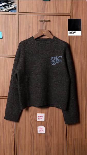 This image ad showcases a dark gray sweater with a light blue monogram, presented in a visually curated setting with col