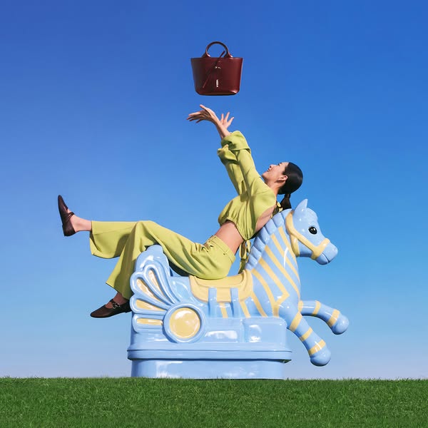 A lifestyle image ad featuring a person on a children's rocking horse reaching up toward a handbag.