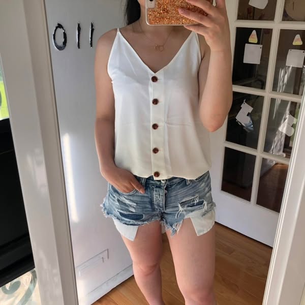 A woman takes a mirror selfie wearing a white button-down tank top and denim shorts.