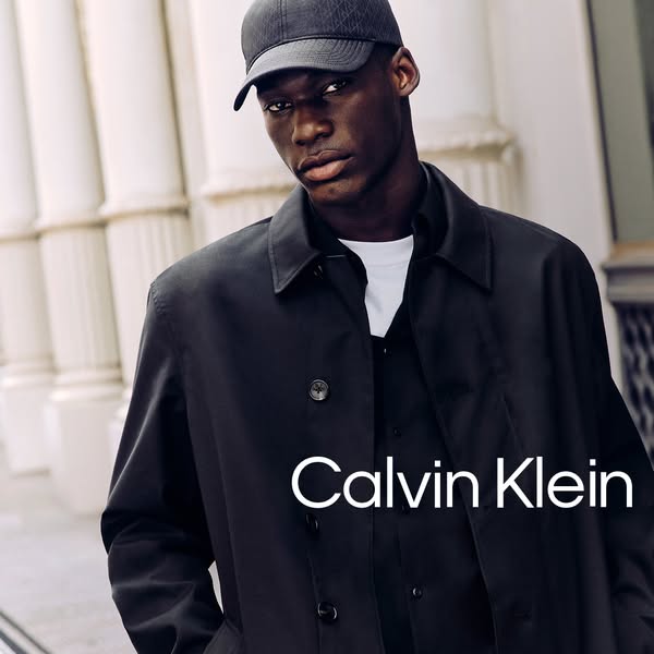 An image ad showcasing the Calvin Klein brand featuring a model wearing a black trench coat and baseball cap against a w