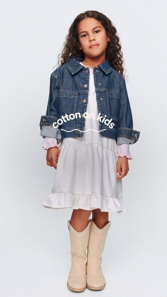 This ad features a young girl modeling clothing from Cotton On Kids. She is wearing a denim jacket, white dress, and boo