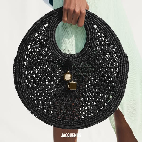 This image ad showcases a black straw handbag by the brand Jacquemus. The bag is held up by a model, highlighting its un