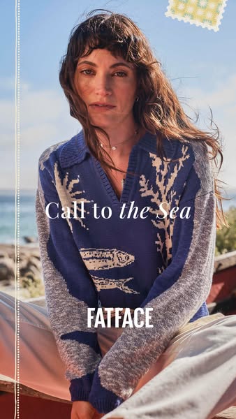 An image of a woman sitting in front of a coastal scene. She is wearing a blue knit sweater with an ocean life motif. Th