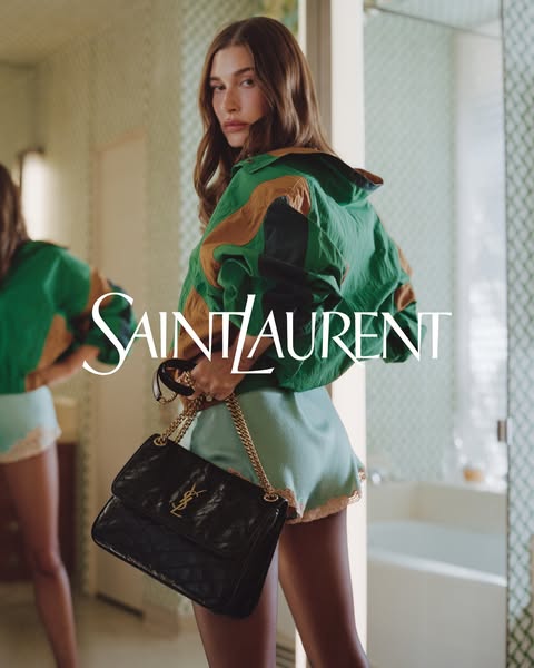 An image ad featuring Hailey Bieber holding a Saint Laurent purse in a bathroom.