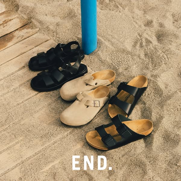 This ad features four pairs of Birkenstock shoes displayed on a sandy surface, with a blue object in the background. The