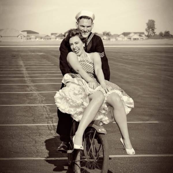 A vintage-style sepia-toned image shows a young woman in a polka dot dress sitting sidesaddle on a motorcycle while a ma