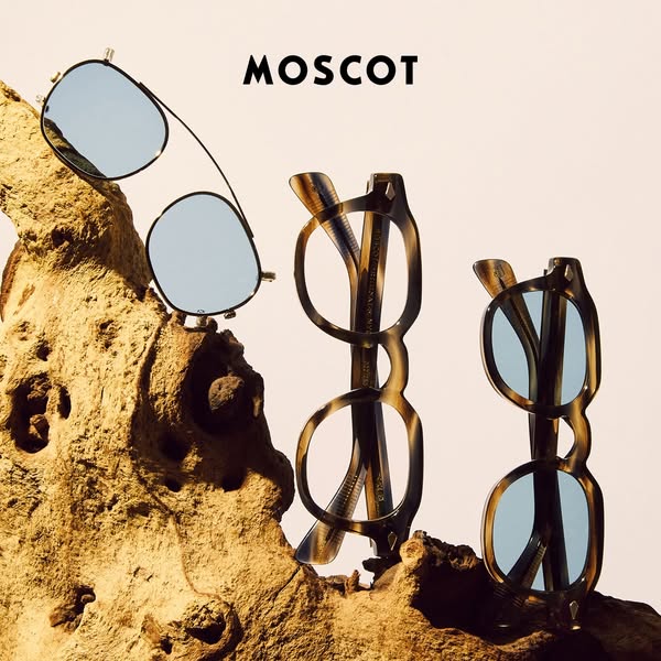 This image ad showcases three MOSCOT eyewear products. A pair of clip-on sunglasses sits atop a wooden display, with two