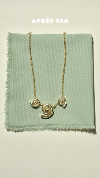 This ad features a gold necklace with spiral pendants against a light green textured fabric. The overall aesthetic is cl