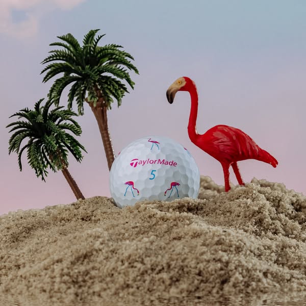 The ad showcases a TaylorMade golf ball set in a miniature scene with sand, palm trees, and a flamingo.
