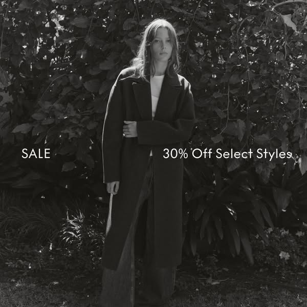 This black and white ad features a woman wearing a long overcoat, paired with jeans and a white shirt, in a natural outd