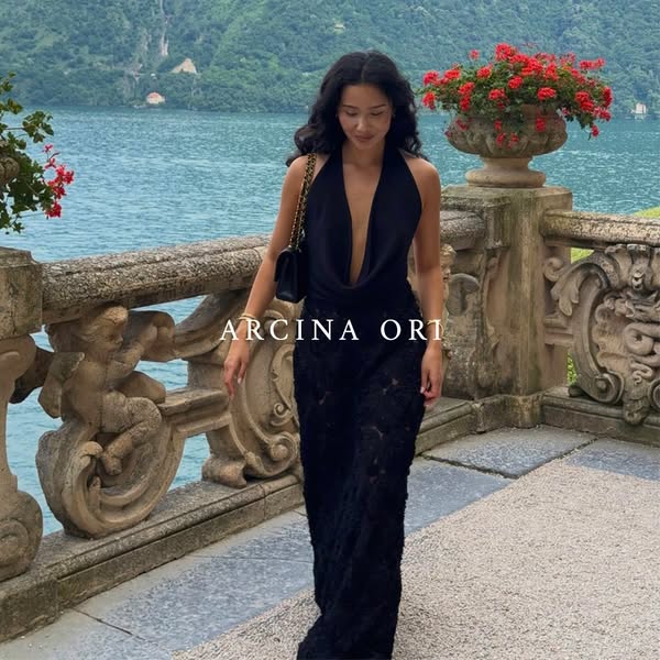 This image ad showcases a woman wearing a black top and pants from Arcina Ori while standing outside with a lake and mou