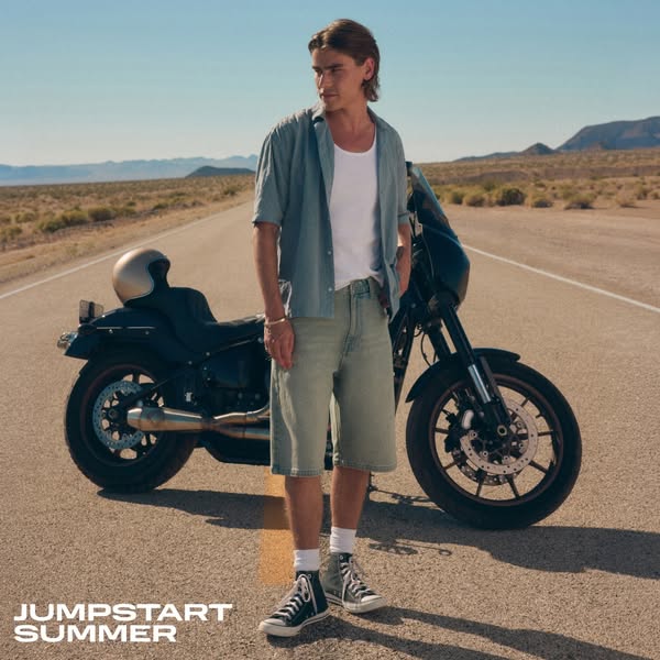 This ad showcases a man in denim shorts standing next to a motorcycle on a desert highway. The ad uses the text "JU