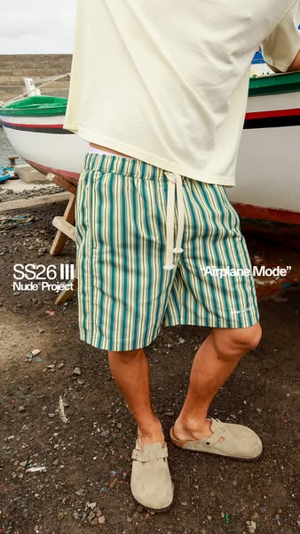 This image ad shows a man wearing Nude Project striped shorts, styled with a beige shirt and matching clogs. The setting