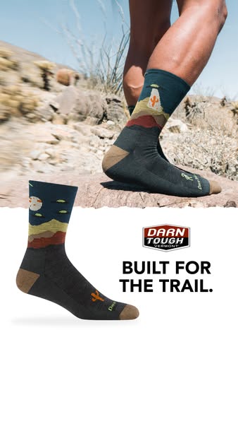 This ad features two Darn Tough socks in a desert landscape setting. The ad emphasizes the socks are built for the trail
