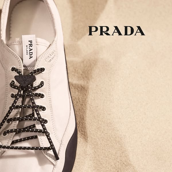 This ad showcases a close-up of a Prada sneaker on a sand-colored surface, emphasizing the brand's luxury and design