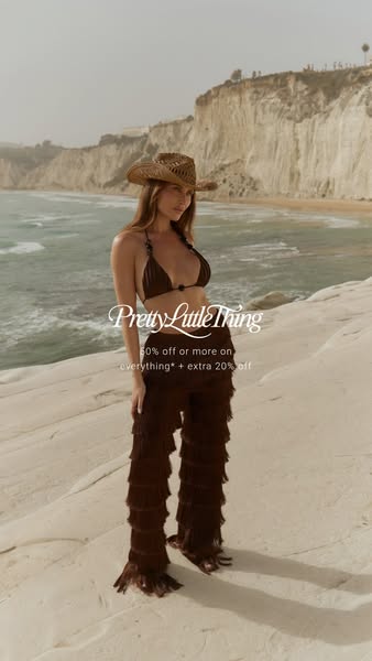 A woman stands in front of a beach and cliff background wearing a bikini and pants.