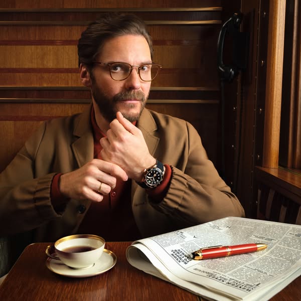 This ad shows a man sitting at a table wearing a watch. A cup of coffee, pen, and newspaper are also on the table. The a