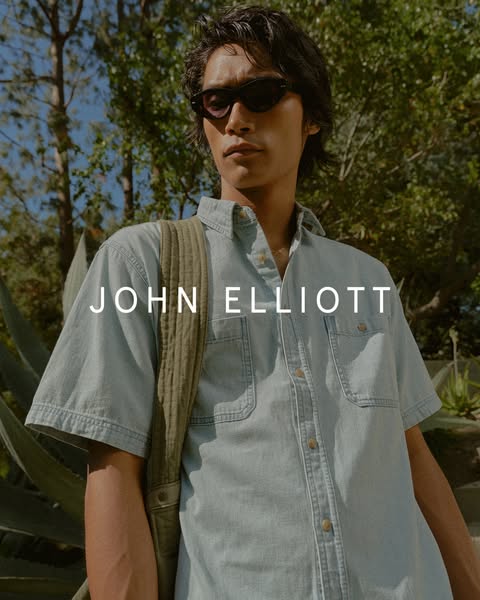 This image ad showcases a male model wearing a John Elliott short-sleeved shirt and sunglasses, with a quilted bag over 