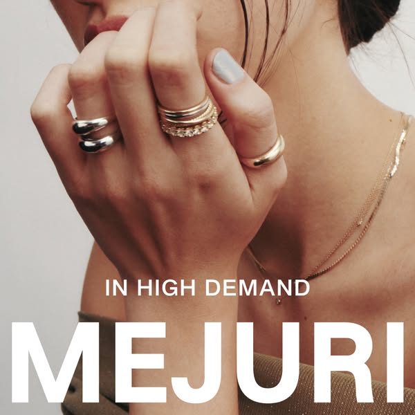 Close up of a model's hand with Mejuri rings and a necklace. The text highlights the brand name and 'in high dem