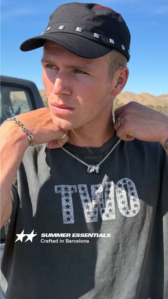 A close up shot of a fair skinned man wearing a Summer Essentials necklace. He is wearing a dark t-shirt, a studded cap,