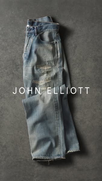 This image ad showcases a pair of distressed denim jeans from the brand John Elliott. The jeans are laid flat on a textu