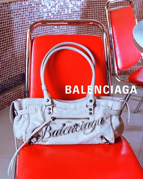 This ad showcases a Balenciaga handbag placed on a red chair in what appears to be a diner setting. The focus is on the 