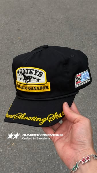 This image ad features a black baseball cap with gold embroidery and various patches. It is being held by a hand that is