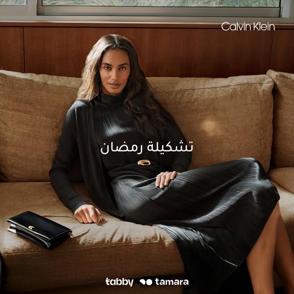 This image ad for Calvin Klein features a woman wearing a pleated black dress and cardigan, sitting on a couch with a bl
