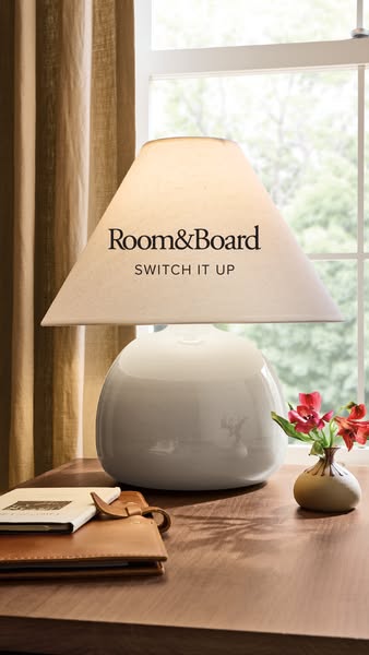 An ad featuring a white lamp from Room & Board on a table in front of a window. The ad highlights the ability to swi