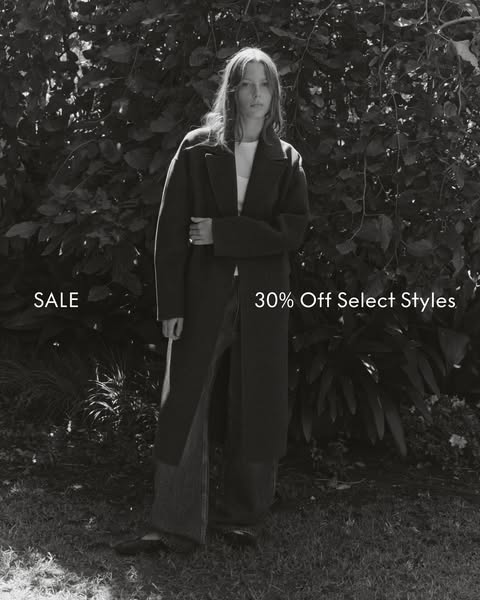 A black and white image ad promoting a sale on select styles. The ad features a woman in a long coat, standing outdoors.