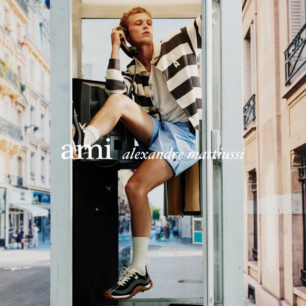 The image shows a young man in a phone booth in Paris, wearing various apparel and footwear items from AMI Paris.