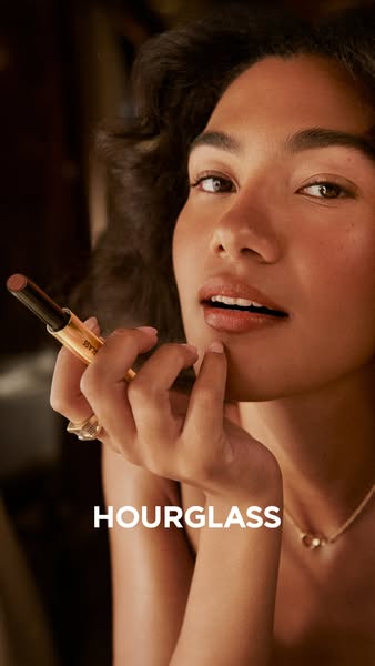 An up-close image focuses on a woman applying Hourglass Phantom Volumizing Glossy Balm. The shot is set at an angle and 