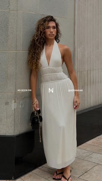 A photo featuring a model wearing a white dress while leaning on a wall and holding a small black handbag. The dress is 