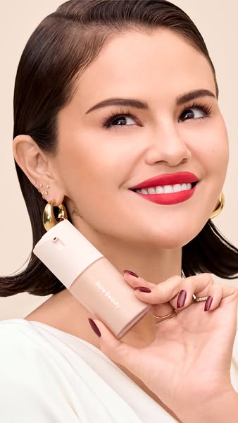 This image promotes Rare Beauty foundation. The image features Selena Gomez holding the product and facing towards the c